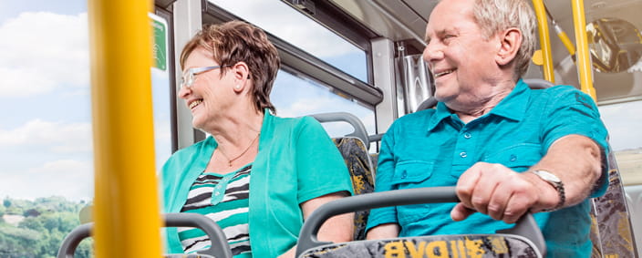 Das Foto zeigt einen Mann und eine Frau, welche im Bus sitzen und lächelnd aus dem Fenster schauen.