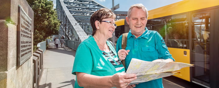 Zdjęcie pokazuje dwóch turystów z planem miasta przed mostem Blaues Wunder, w tle autobus DVB
