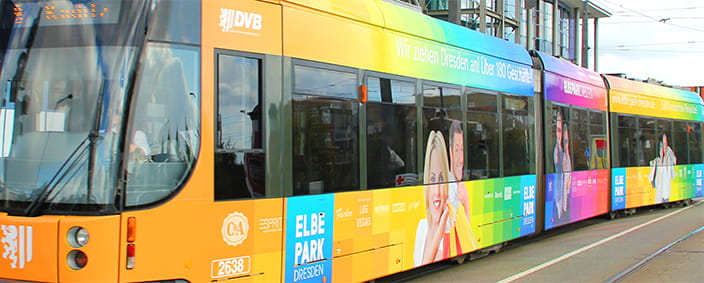 Das Bild zeigt eine Straßenbahn in Regenbogenfarben mit Werbung des Dresdner Elbe Parks