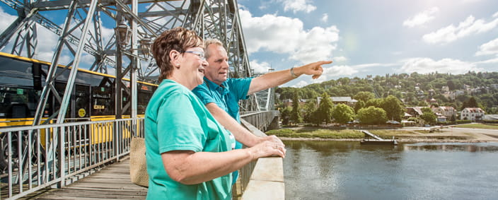 The photo shows two tourists looking down at the Elbe from the Blue Wonder bridge. In the background is the Loschwitz hillside.