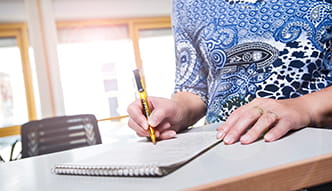 Das Foto zeigt eine Frau, welche an einem Tisch sitzt und auf einem Block schreibt.